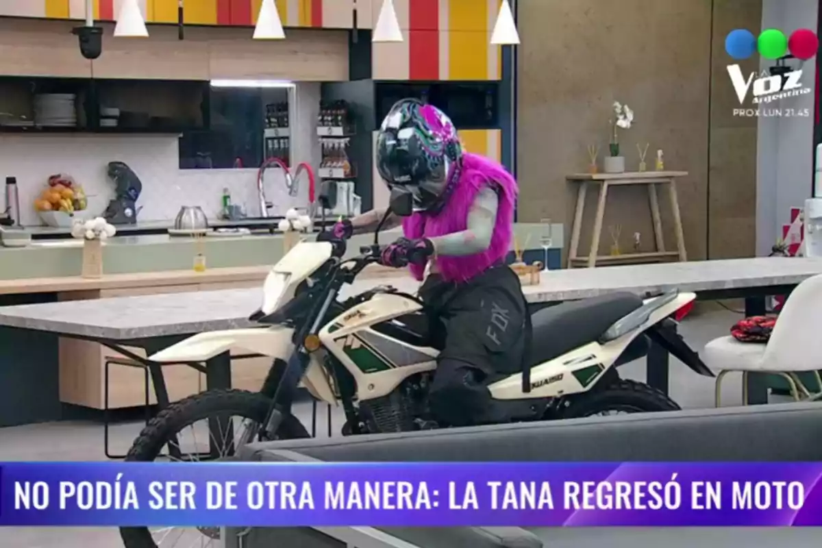 Persona con casco y chaleco rosa conduciendo una moto dentro de una cocina moderna mientras en la parte inferior aparece un texto en español Persona con casco y chaleco rosa conduciendo una moto dentro de una cocina moderna mientras en la parte inferior aparece un texto en español