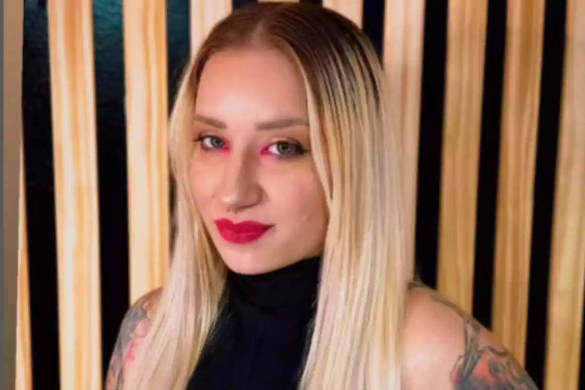 Mujer de cabello rubio largo y lacio con labios rojos y maquillaje llamativo, usando blusa negra sin mangas y fondo de listones de madera verticales