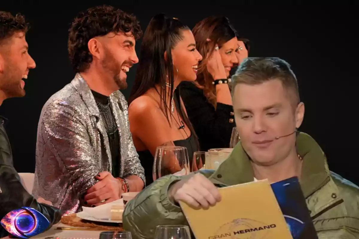 A group of people smile and talk at a table while a man with a microphone holds a yellow Big Brother envelope.