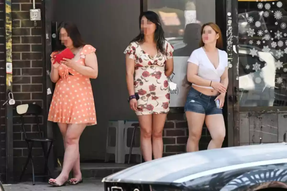Three women with pixelated faces are standing on the sidewalk in front of a brick building, each wearing summer clothes and relaxed expressions. Three women with pixelated faces are standing on the sidewalk in front of a brick building, each wearing summer clothes and relaxed expressions.