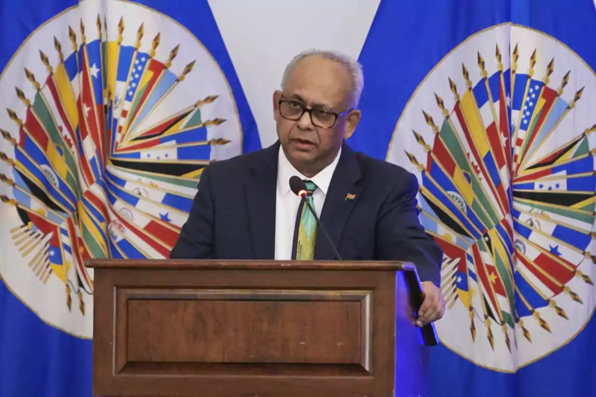 Hombre de traje hablando en un podio con dos banderas coloridas de fondo