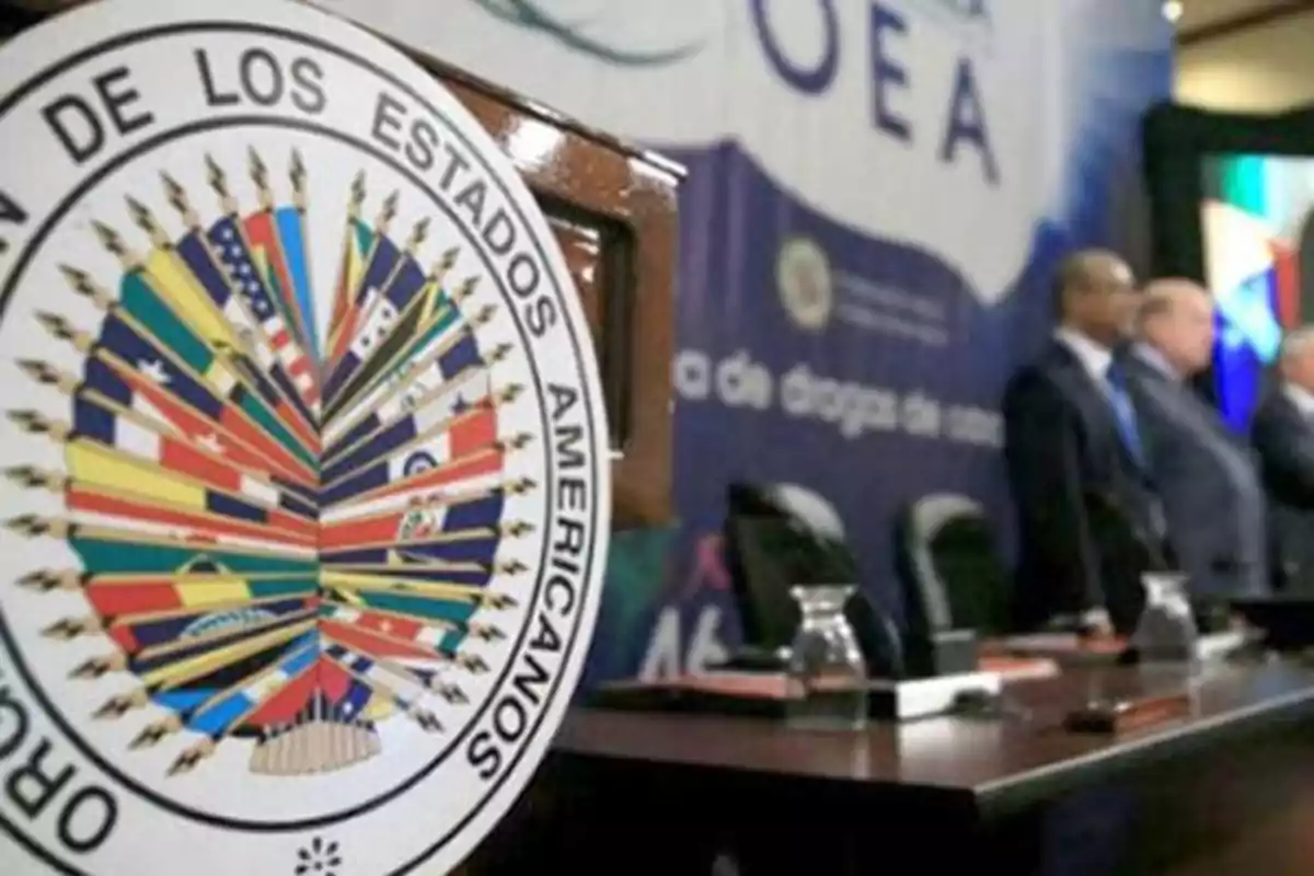 Escudo de la Organización de los Estados Americanos en primer plano con una mesa de conferencia y personas al fondo