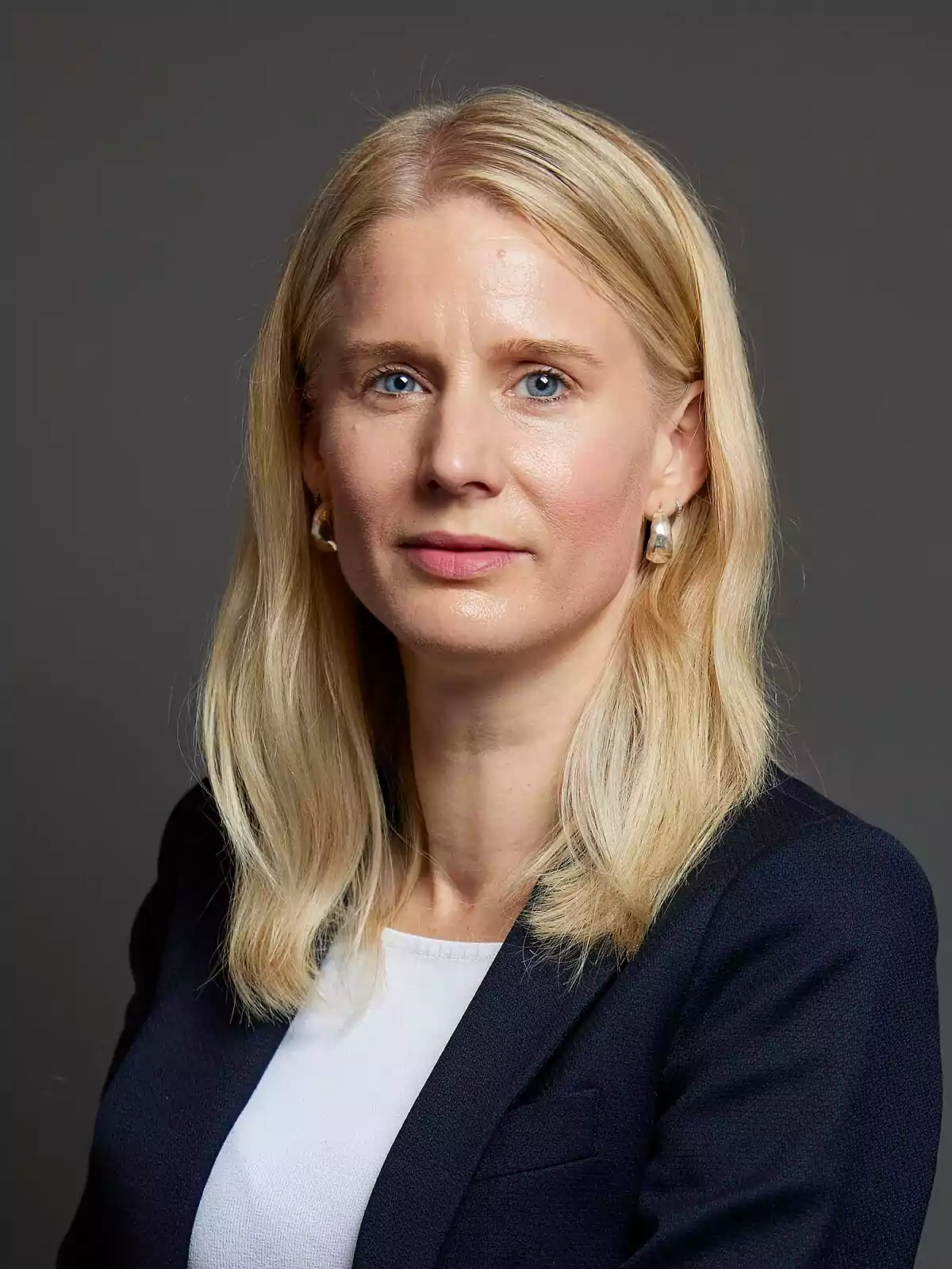 Blonde woman with straight, loose hair wearing a navy blue jacket and a white blouse posing in front of a gray background Blonde woman with straight, loose hair wearing a navy blue jacket and a white blouse posing in front of a gray background