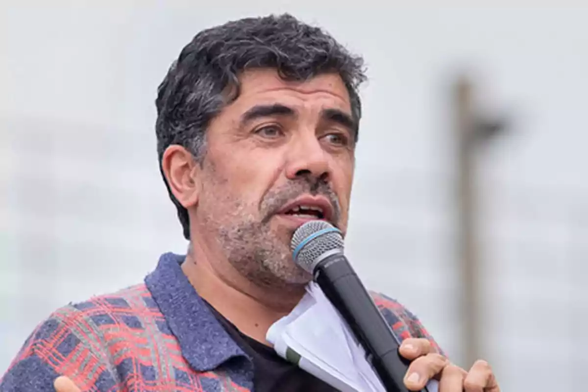Hombre de cabello oscuro y barba hablando por un micrófono mientras sostiene unos papeles