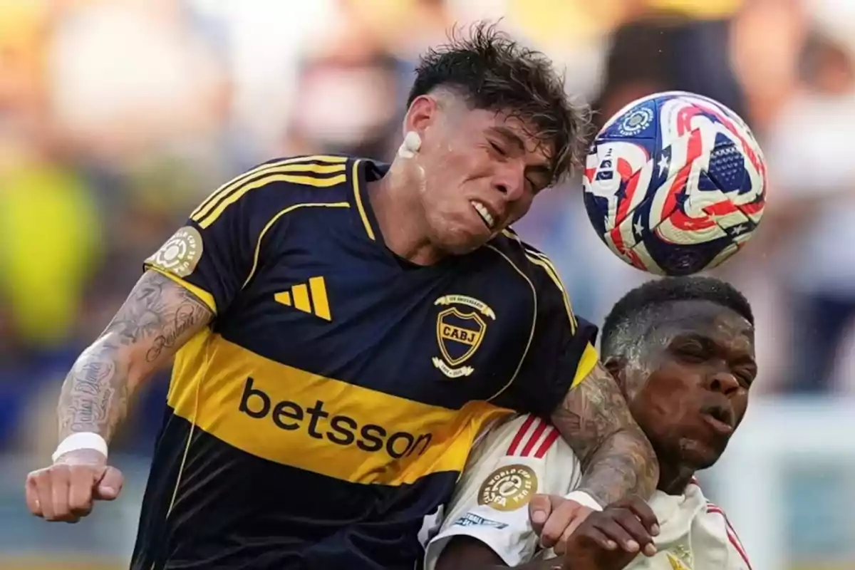 Dos futbolistas disputan un balón en el aire durante un partido, uno de ellos con camiseta azul y amarilla y el otro con camiseta blanca, ambos con expresiones de esfuerzo. Dos futbolistas disputan un balón en el aire durante un partido, uno de ellos con camiseta azul y amarilla y el otro con camiseta blanca, ambos con expresiones de esfuerzo.