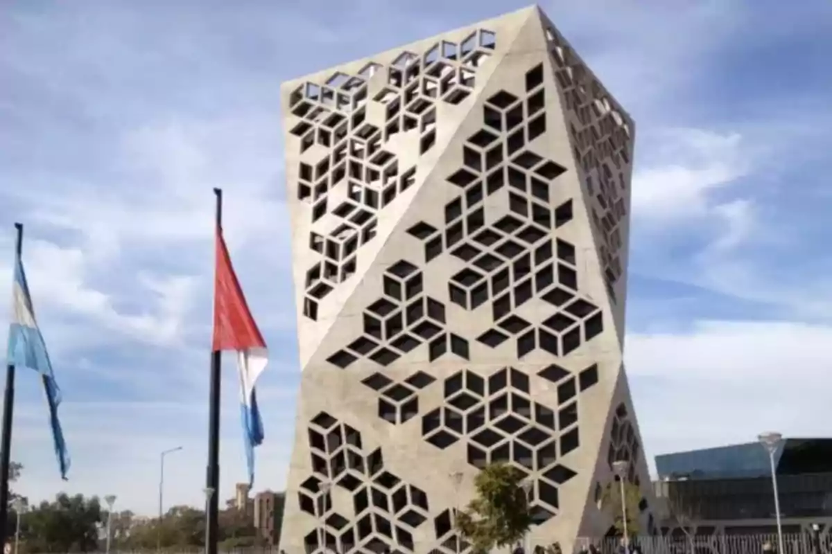 Edificio moderno con diseño geométrico en la fachada y dos banderas ondeando al frente bajo un cielo parcialmente nublado