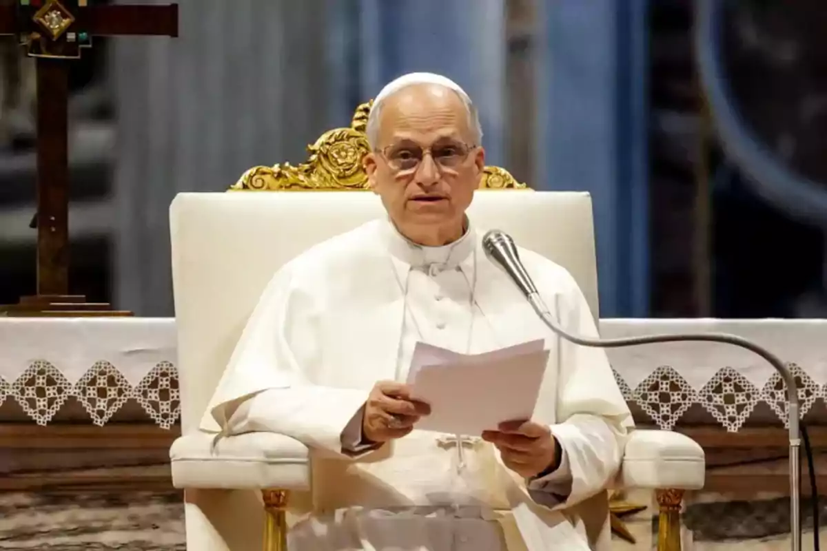 Man dressed in a white robe and zucchetto sitting on a golden armchair, holding papers and speaking into a microphone in a religious setting