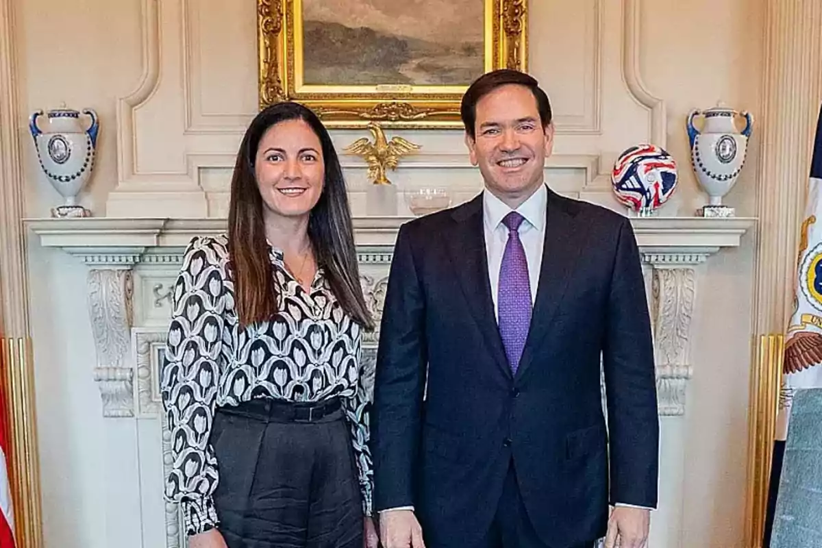 Two people pose smiling in an elegant room decorated with vases, a flag, and a painting on the wall.