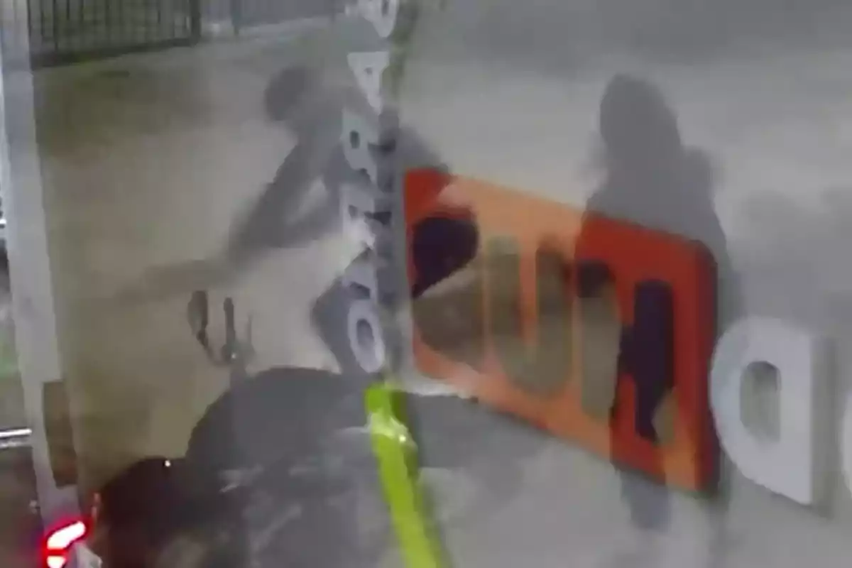 Imagen borrosa de una cámara de seguridad donde se observan dos sombras humanas y una motocicleta junto a un letrero reflejado en una superficie. Imagen borrosa de una cámara de seguridad donde se observan dos sombras humanas y una motocicleta junto a un letrero reflejado en una superficie.