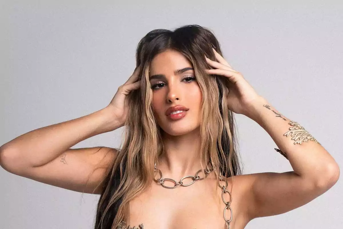 Mujer joven con cabello largo y rubio oscuro, usando un collar de eslabones grandes y tocándose la cabeza con ambas manos, fondo gris claro.
