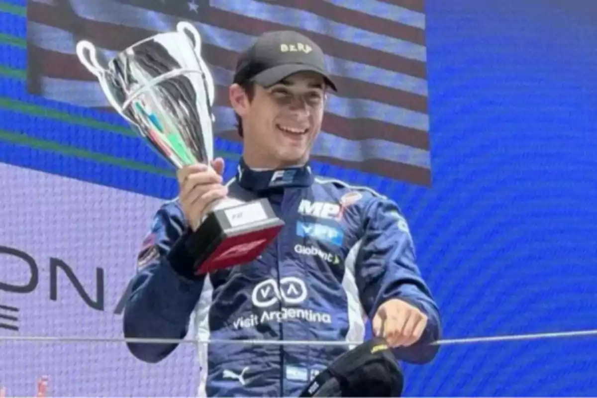 Piloto de automovilismo sonriente sostiene un trofeo en el podio con un uniforme azul y una gorra negra mientras una bandera ondea de fondo Piloto de automovilismo sonriente sostiene un trofeo en el podio con un uniforme azul y una gorra negra mientras una bandera ondea de fondo