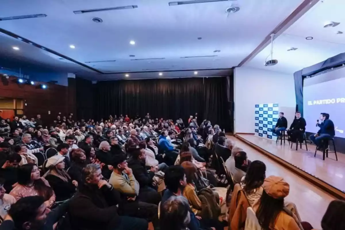Auditorio lleno de personas escuchando a tres oradores sentados en el escenario durante una conferencia o charla Auditorio lleno de personas escuchando a tres oradores sentados en el escenario durante una conferencia o charla