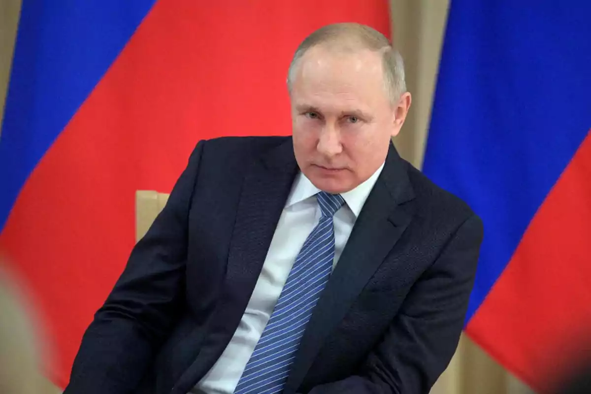 Man in a dark suit and blue tie sitting in front of red and blue flags