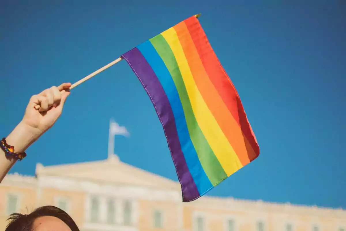 Person holding an LGBT pride flag with a building and blue sky in the background Person holding an LGBT pride flag with a building and blue sky in the background