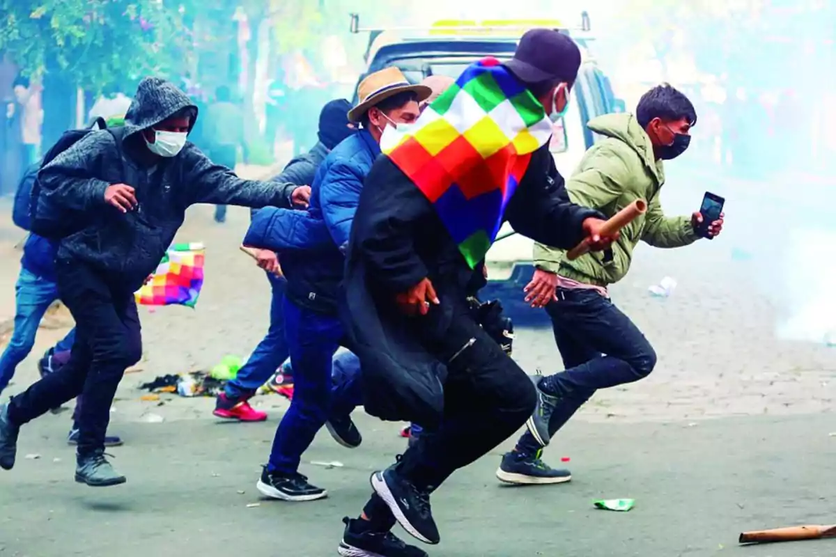 Personas corriendo en una calle durante una protesta, algunas llevan mascarillas y una bandera multicolor, el ambiente está lleno de humo