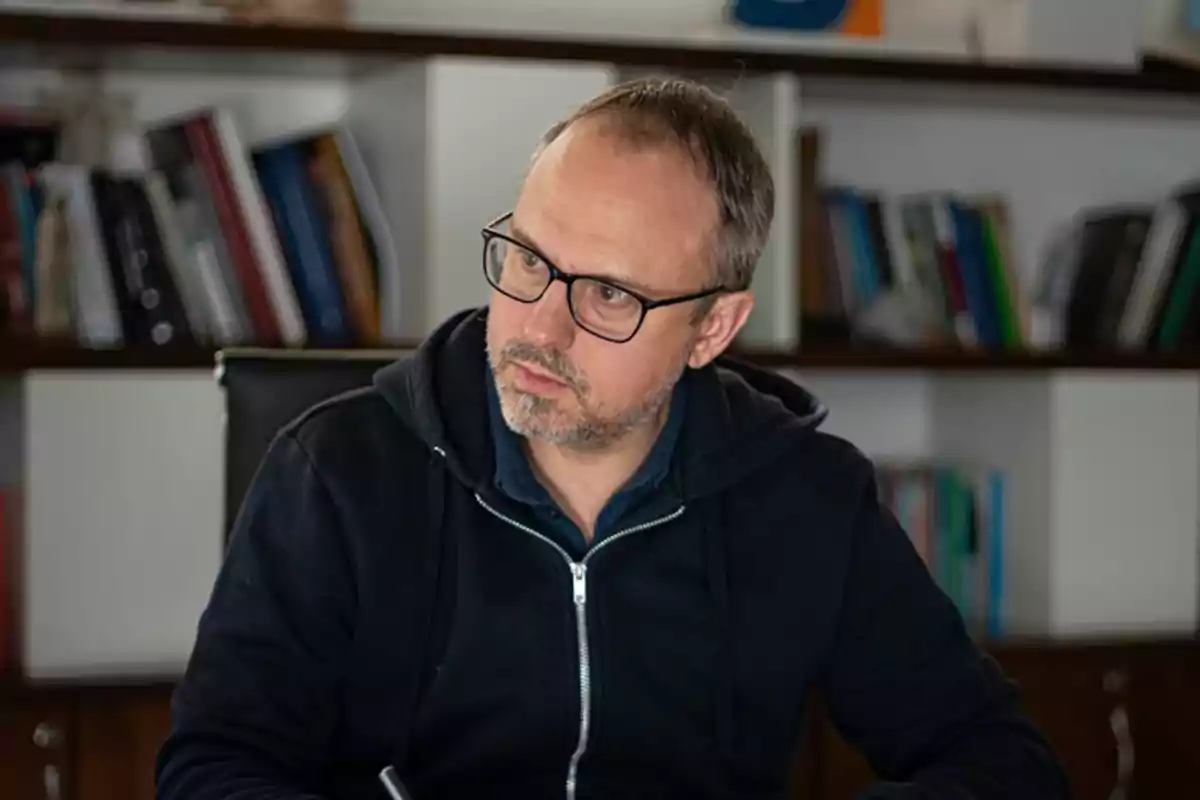 Man with glasses and a short beard sitting in an office with bookshelves full of books in the background Man with glasses and a short beard sitting in an office with bookshelves full of books in the background