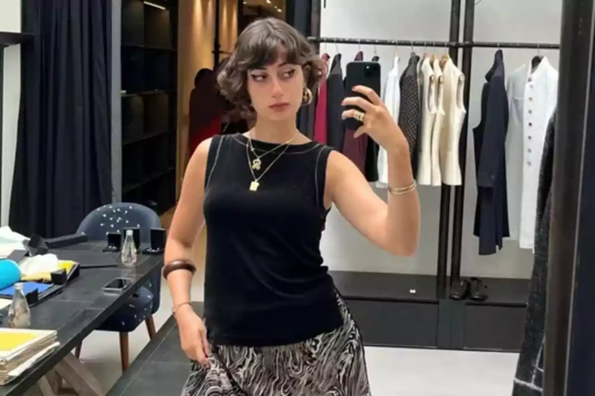 Mujer joven con cabello corto y rizado toma una selfie frente a un espejo en una tienda de ropa elegante, lleva un top negro sin mangas y falda estampada, al fondo se ven prendas colgadas y una mesa con objetos.