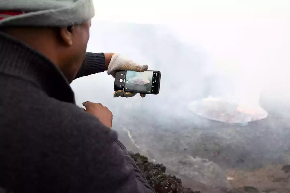 Persona con guantes toma una foto con su celular de un volcán activo cubierto de humo