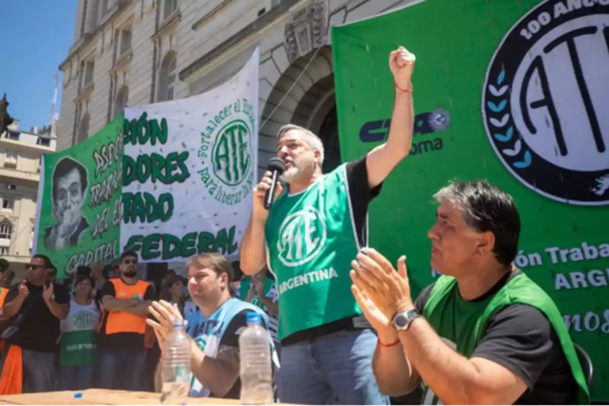 Un grupo de personas participa en una manifestación sindical frente a un edificio, con pancartas y chalecos verdes de la Asociación de Trabajadores del Estado, mientras un hombre habla por micrófono y levanta el puño