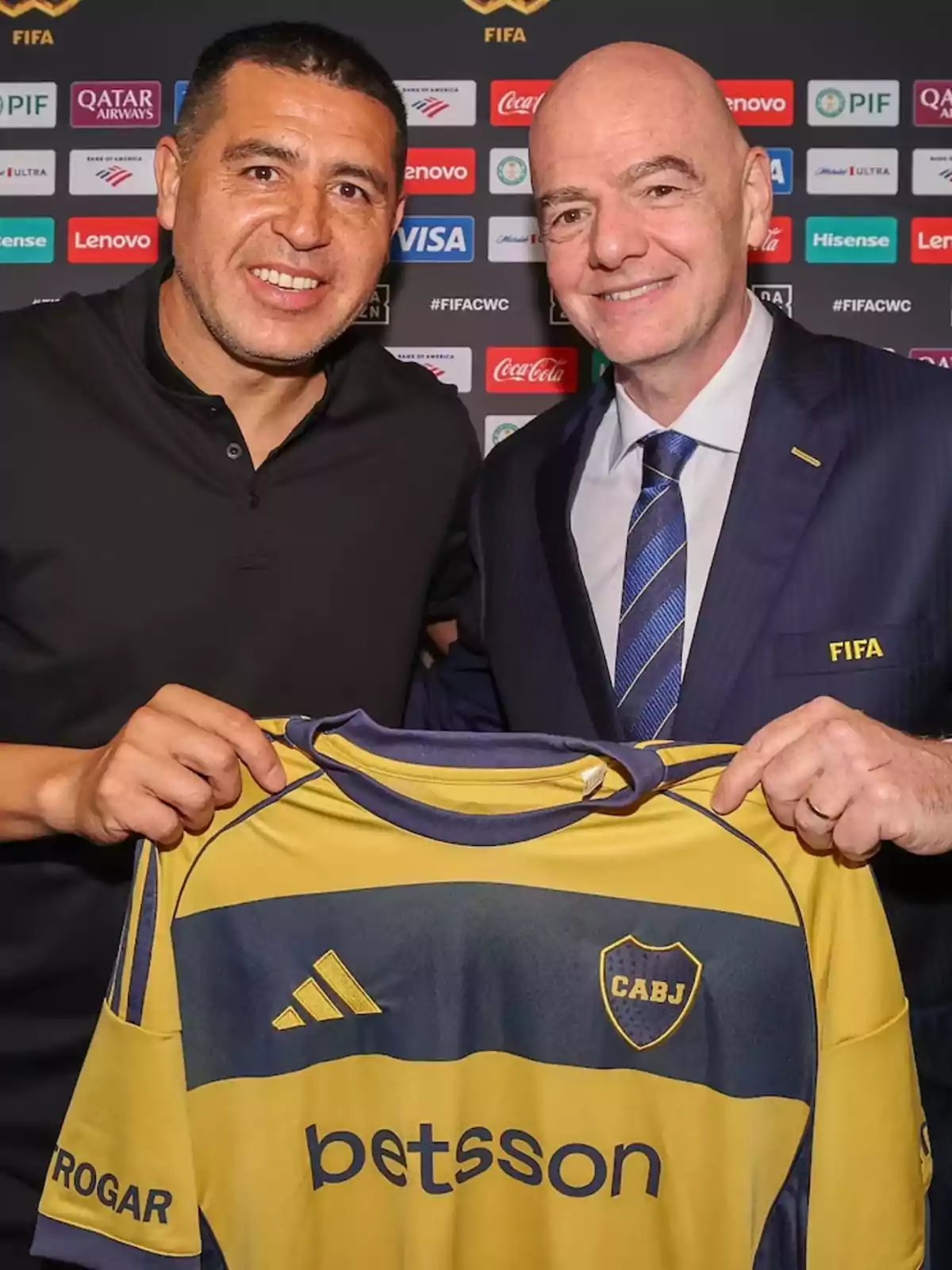 Dos hombres sonríen mientras sostienen una camiseta de Boca Juniors frente a un fondo con logotipos de patrocinadores y FIFA Dos hombres sonríen mientras sostienen una camiseta de Boca Juniors frente a un fondo con logotipos de patrocinadores y FIFA
