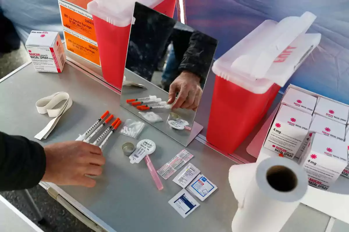 A hand picks up a syringe from a table with medical supplies such as a tourniquet, wipes, boxes of Narcan, a mirror, and red sharps containers. A hand picks up a syringe from a table with medical supplies such as a tourniquet, wipes, boxes of Narcan, a mirror, and red sharps containers.