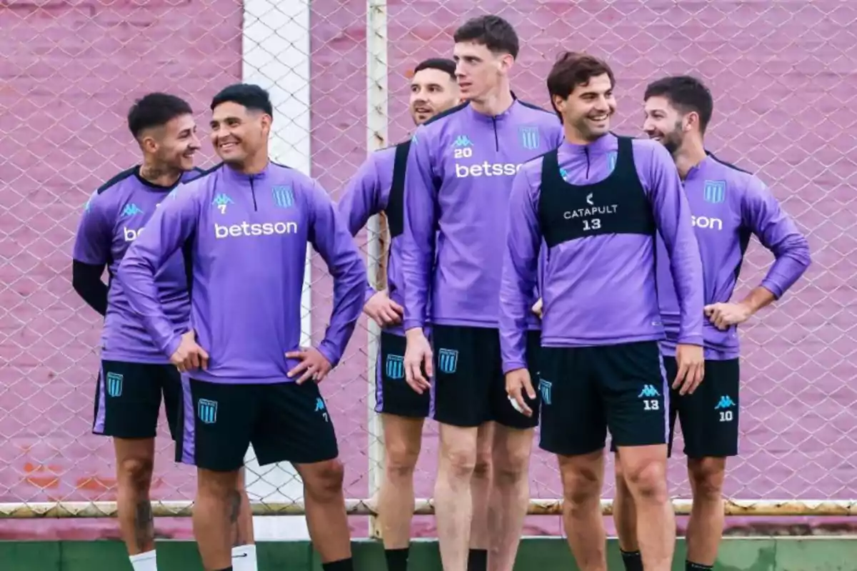 Seis jugadores de fútbol con uniforme de entrenamiento violeta posan sonrientes frente a una reja en una cancha