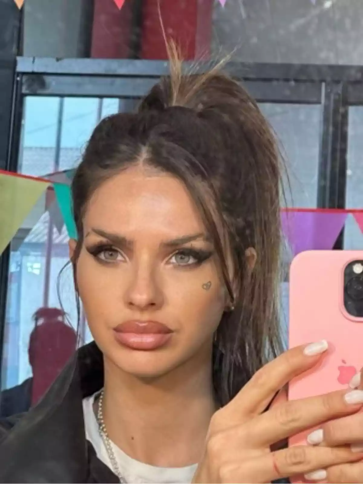 Mujer con cabello largo y oscuro recogido en una coleta alta, maquillaje marcado y un pequeño tatuaje de corazón en la mejilla, tomándose una selfie con un teléfono móvil rosa frente a un espejo decorado con banderines de colores. Mujer con cabello largo y oscuro recogido en una coleta alta, maquillaje marcado y un pequeño tatuaje de corazón en la mejilla, tomándose una selfie con un teléfono móvil rosa frente a un espejo decorado con banderines de colores.
