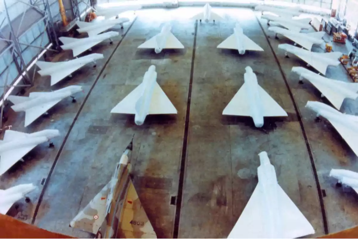 Varios aviones de combate con alas en delta alineados dentro de un hangar, la mayoría cubiertos con lonas blancas y uno descubierto en primer plano. Varios aviones de combate con alas en delta alineados dentro de un hangar, la mayoría cubiertos con lonas blancas y uno descubierto en primer plano.