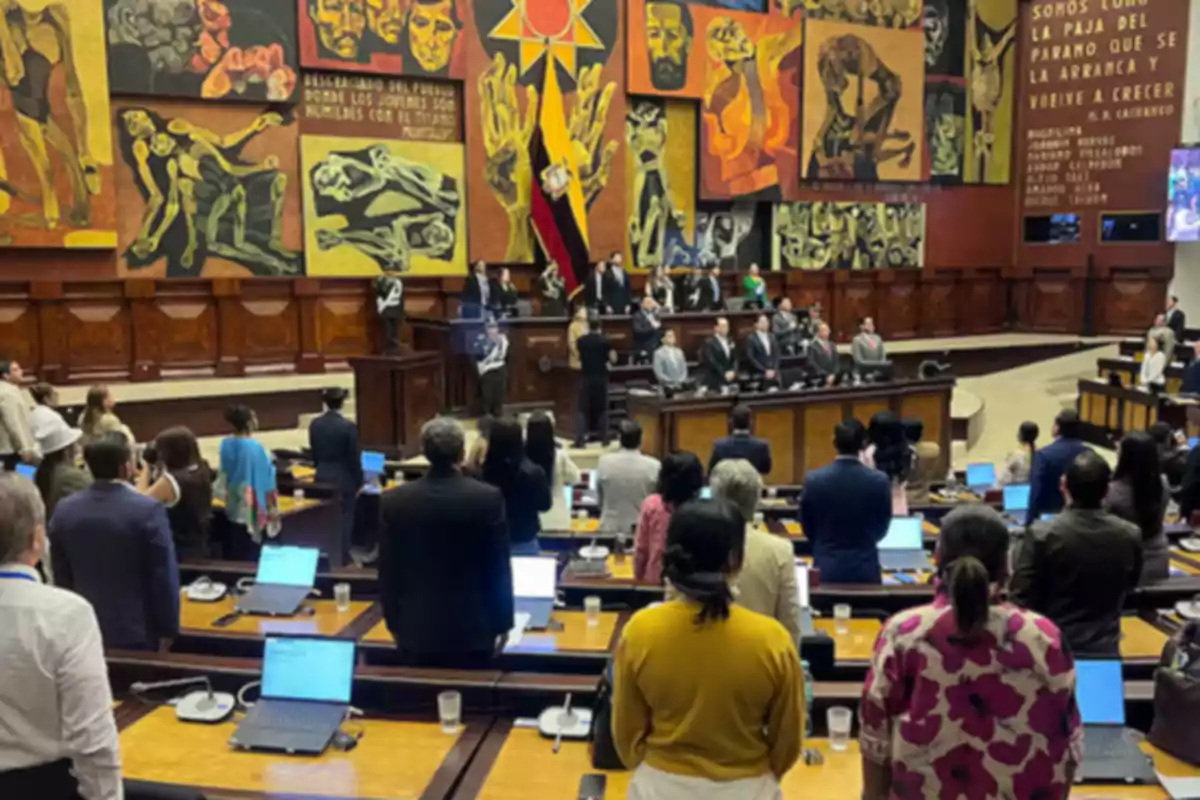 Personas reunidas en el pleno de la Asamblea Nacional de Ecuador con murales coloridos en las paredes y la bandera nacional al fondo