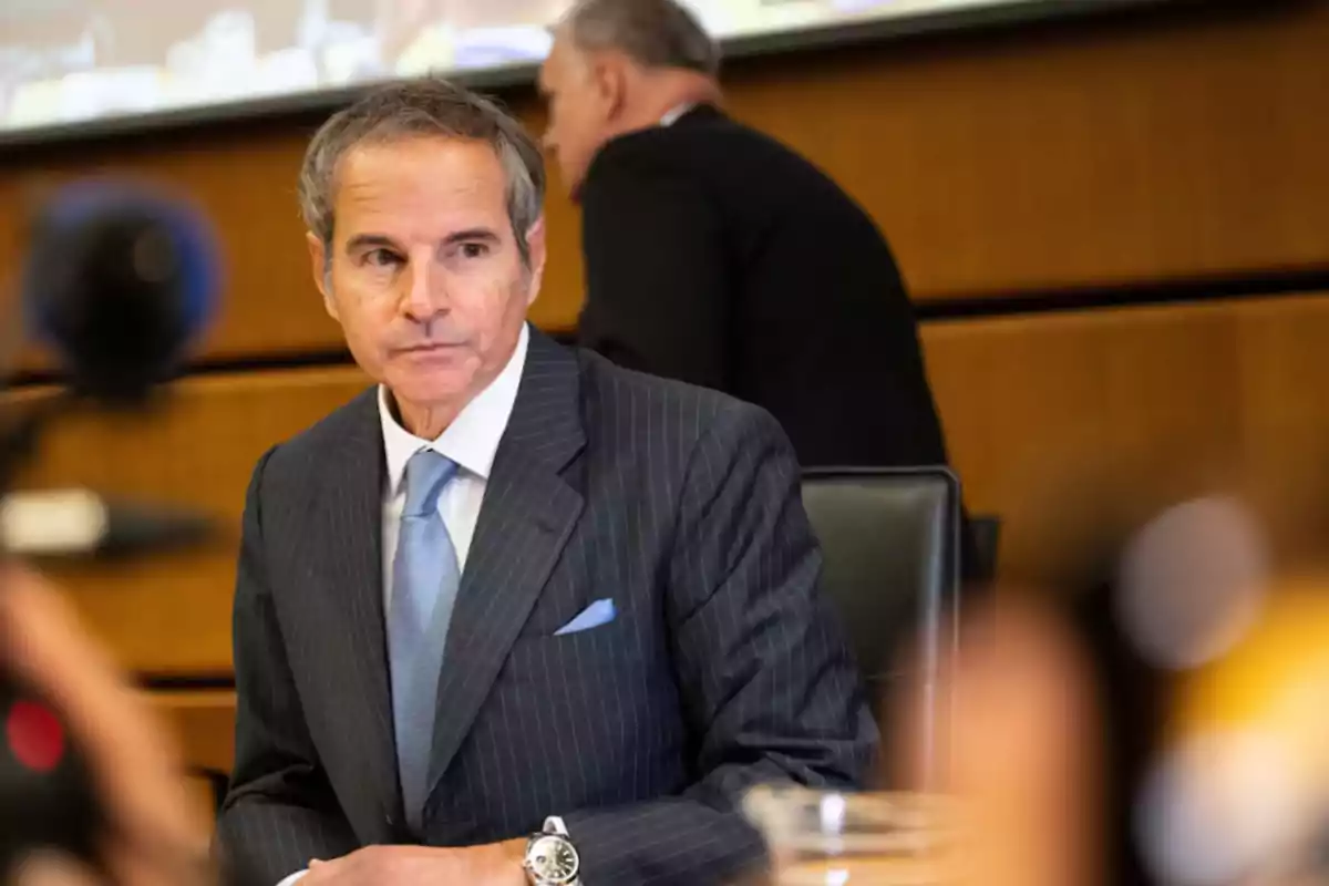 Hombre de traje gris con corbata azul claro sentado en una sala de conferencias mirando hacia un lado mientras otra persona pasa detrás de él