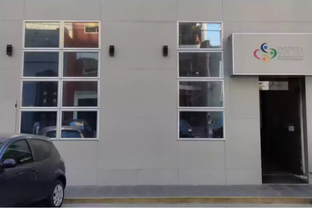 Fachada de un edificio gris con ventanas rectangulares y un cartel de OSPMA junto a una puerta de entrada y un auto estacionado a la izquierda Fachada de un edificio gris con ventanas rectangulares y un cartel de OSPMA junto a una puerta de entrada y un auto estacionado a la izquierda