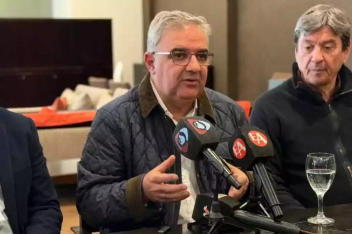 Un hombre de cabello canoso y gafas habla frente a varios micrófonos en una conferencia de prensa, acompañado por otra persona y con una copa de agua sobre la mesa.