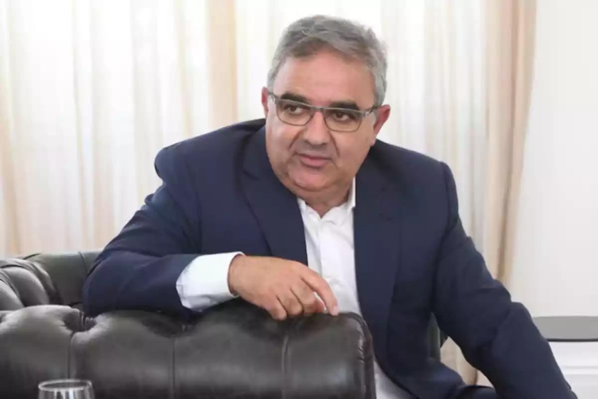 Hombre de cabello canoso y gafas sentado en un sillón de cuero negro vistiendo un traje azul y camisa blanca con cortinas claras de fondo