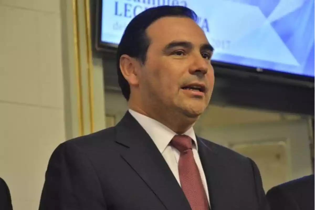 Dark-haired man in a suit and red tie speaking at a formal event