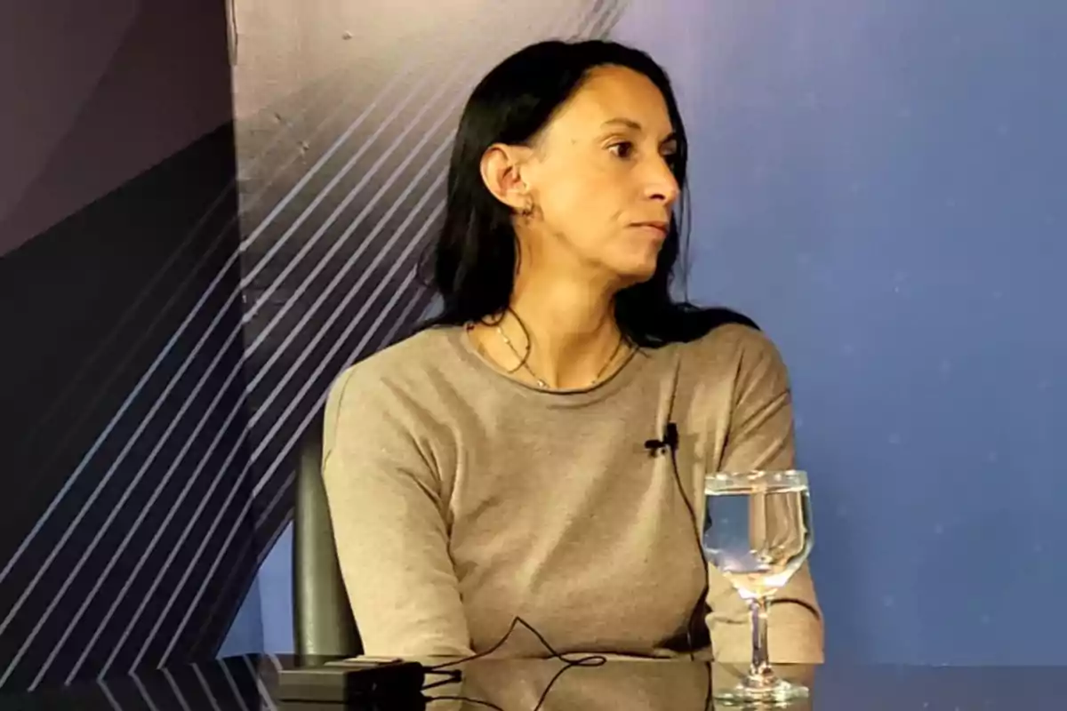 Dark-haired woman sitting at a table with a lapel microphone and a glass of water in front of her in a studio setting Dark-haired woman sitting at a table with a lapel microphone and a glass of water in front of her in a studio setting