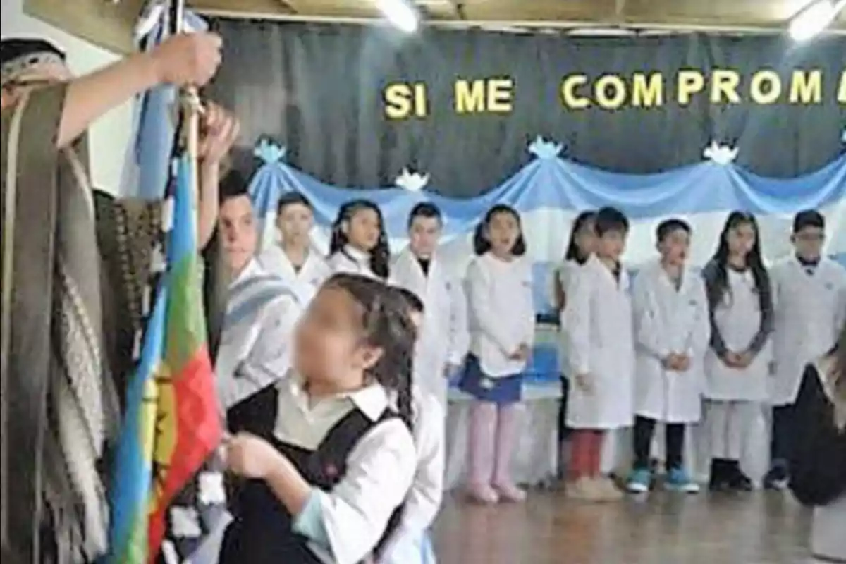 Boys and girls wearing white smocks participate in a school event; in the background, there is an Argentine flag and yellow letters forming an incomplete phrase, while someone holds a Mapuche flag.
