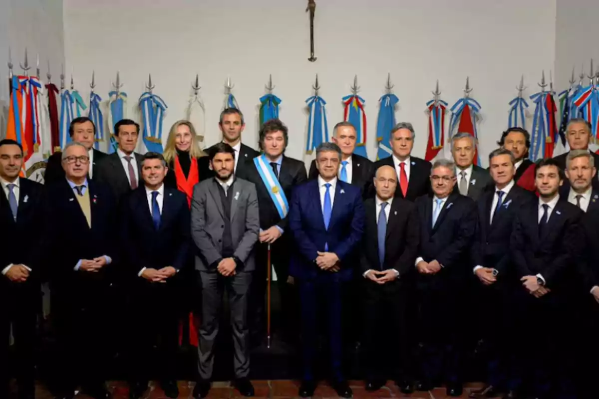Grupo de personas vestidas formalmente posando para una foto oficial frente a banderas argentinas en un salón institucional Grupo de personas vestidas formalmente posando para una foto oficial frente a banderas argentinas en un salón institucional