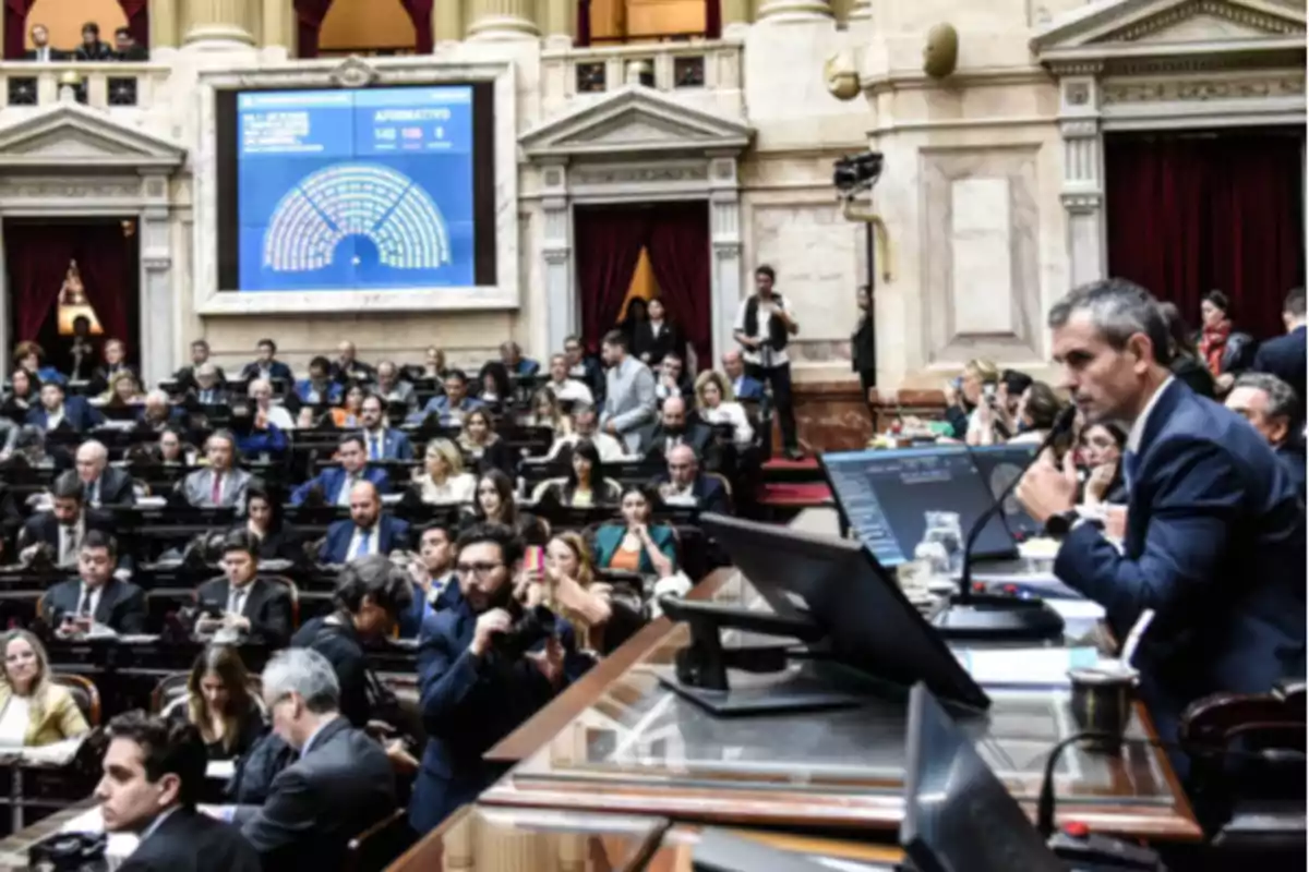 Parliamentary session in a legislative chamber with numerous deputies seated and a speaker of the house speaking into a microphone while the results of a vote are displayed on a large screen. Parliamentary session in a legislative chamber with numerous deputies seated and a speaker of the house speaking into a microphone while the results of a vote are displayed on a large screen.