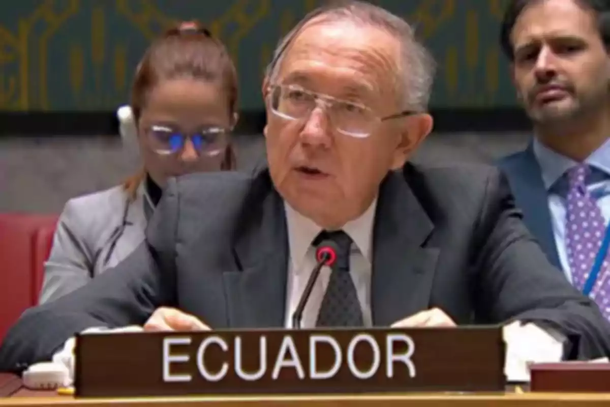 Hombre mayor con gafas y traje hablando en una conferencia internacional con un letrero que dice Ecuador frente a él Hombre mayor con gafas y traje hablando en una conferencia internacional con un letrero que dice Ecuador frente a él
