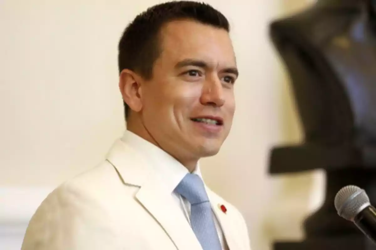 Young man with short dark hair wearing a white suit and light blue tie speaking in front of a microphone Young man with short dark hair wearing a white suit and light blue tie speaking in front of a microphone