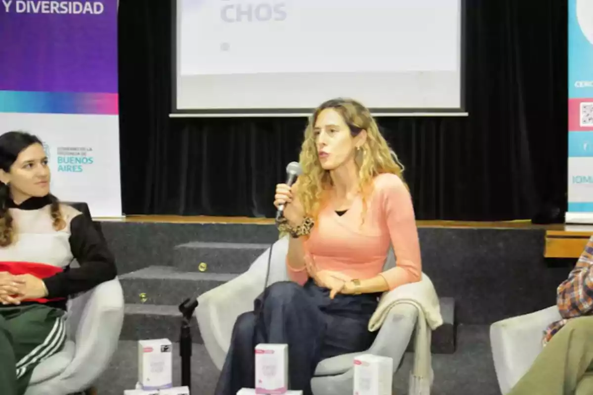 Blonde, wavy-haired woman speaking into a microphone while seated on a panel with others in an auditorium