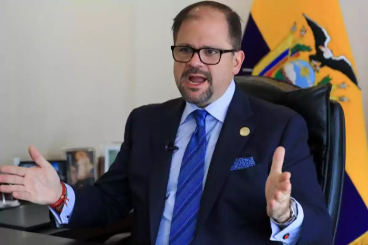 Man in a blue suit gesturing while speaking in an office with an Ecuadorian flag in the background