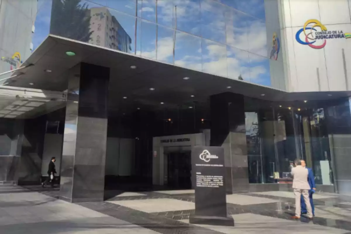 Fachada de un edificio moderno de vidrio con el logo del Consejo de la Judicatura y personas caminando cerca de la entrada principal Fachada de un edificio moderno de vidrio con el logo del Consejo de la Judicatura y personas caminando cerca de la entrada principal