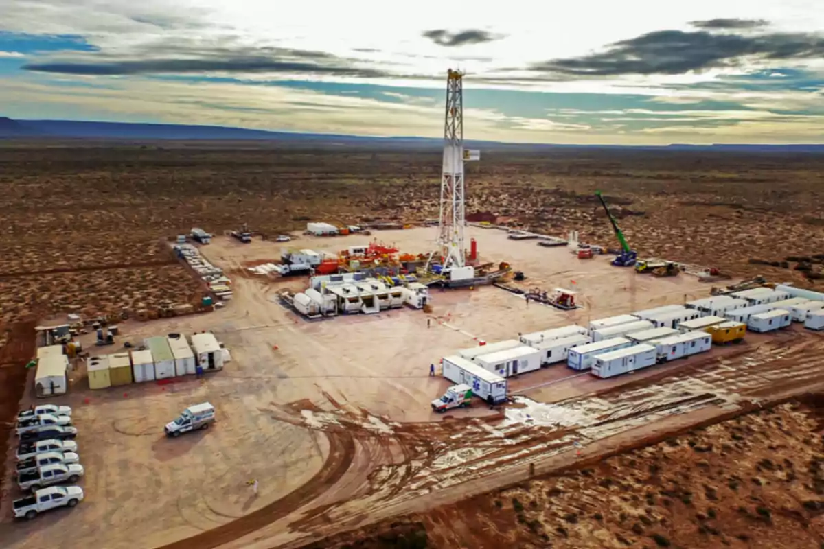 Aerial view of an oil field with a drilling rig surrounded by vehicles, containers, and mobile offices in a desert area.