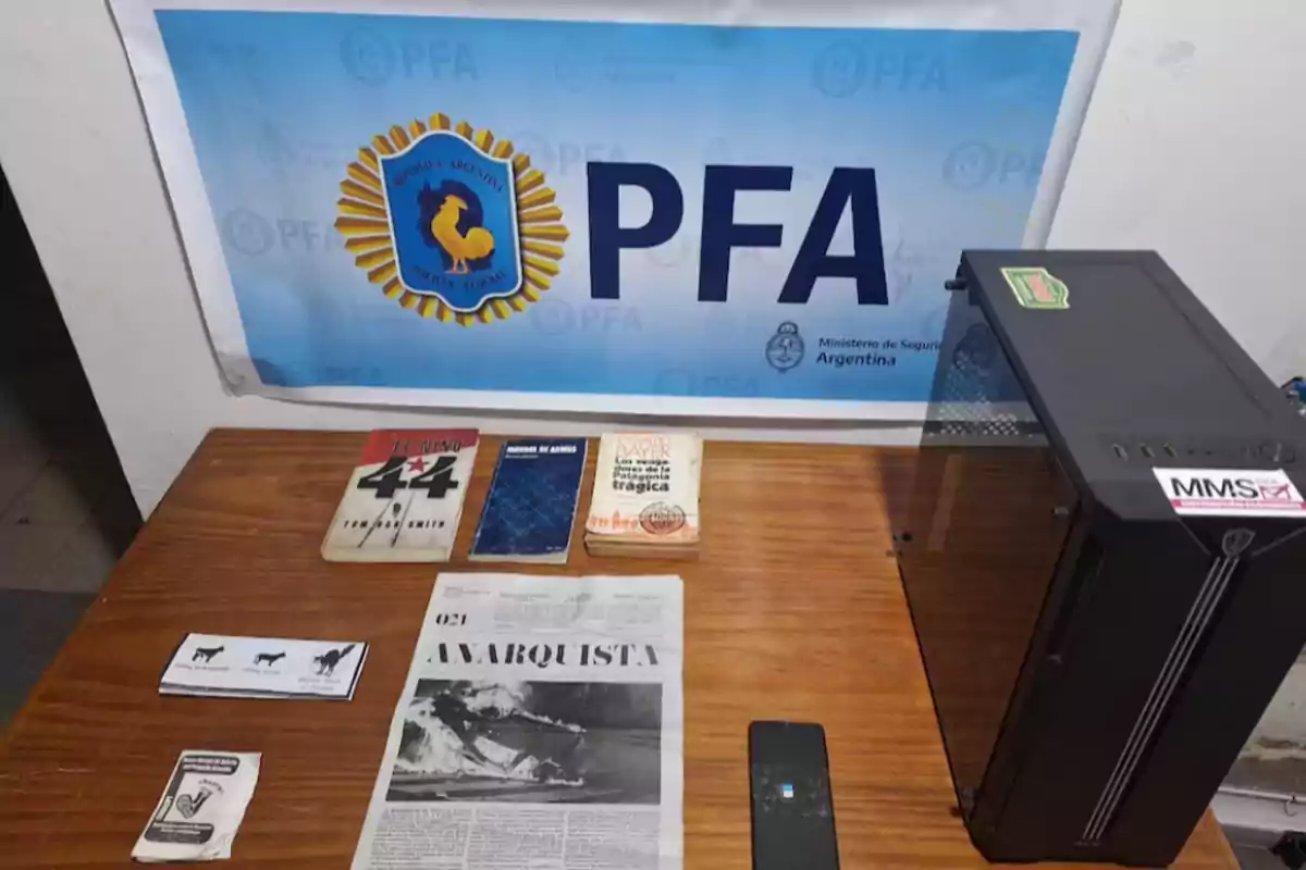 On a wooden table there are books, brochures, a newspaper titled Anarquista, a mobile phone, and a desktop computer next to a poster of the Argentine Federal Police on the back wall. On a wooden table there are books, brochures, a newspaper titled Anarquista, a mobile phone, and a desktop computer next to a poster of the Argentine Federal Police on the back wall.