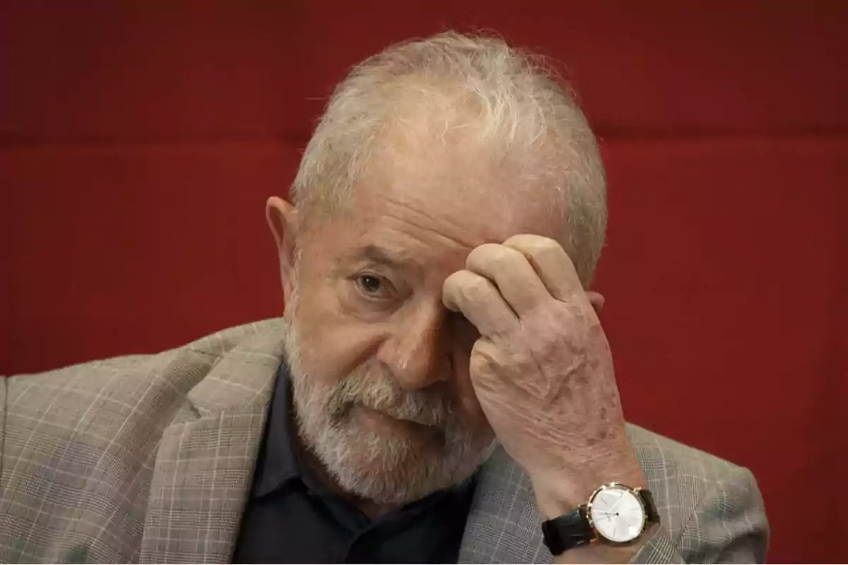 Hombre mayor con barba y cabello canoso vestido con saco gris y camisa oscura tocándose la frente con expresión seria frente a un fondo rojo