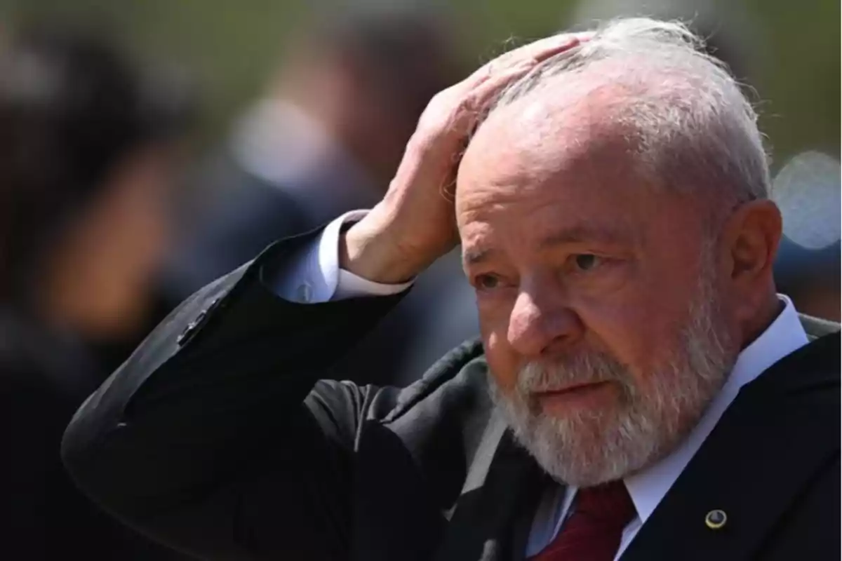 Hombre mayor de barba y cabello canoso vestido con traje oscuro y corbata roja se toca la cabeza con expresión seria