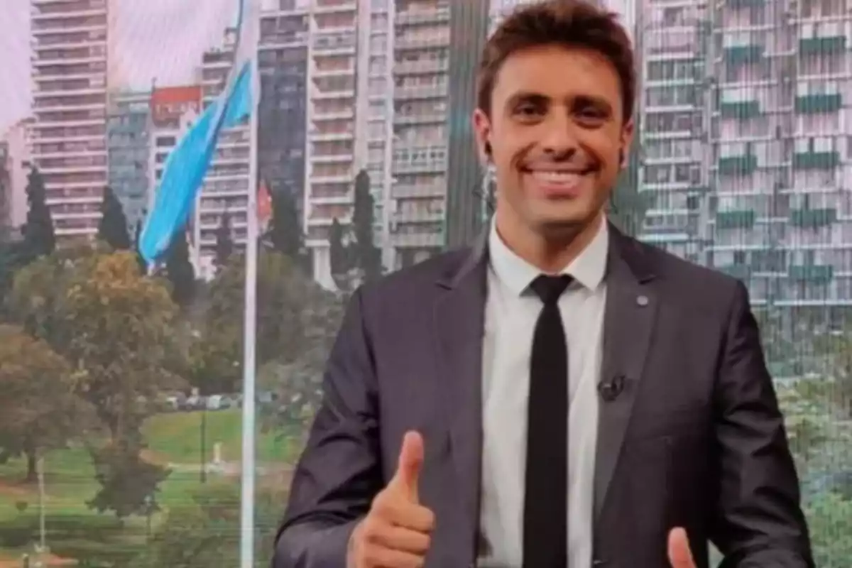 Hombre joven con traje y corbata sonriendo y haciendo el gesto de pulgar arriba frente a un fondo urbano con edificios y una bandera azul y blanca ondeando