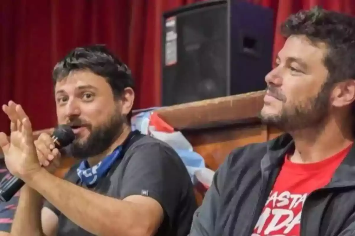 Dos hombres sentados en una mesa, uno de ellos hablando por micrófono mientras el otro lo observa, con fondo de cortinas rojas y una bandera detrás. Dos hombres sentados en una mesa, uno de ellos hablando por micrófono mientras el otro lo observa, con fondo de cortinas rojas y una bandera detrás.