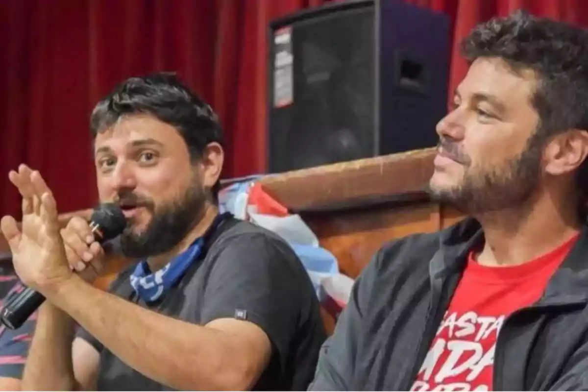 Dos hombres sentados en un escenario, uno de ellos sostiene un micrófono y gesticula mientras el otro lo observa sonriendo, al fondo se ven cortinas rojas y un altavoz.